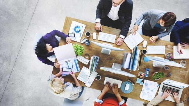 Ein großer Arbeitstisch mit mehreren arbeitenden Menschen daran von Oben