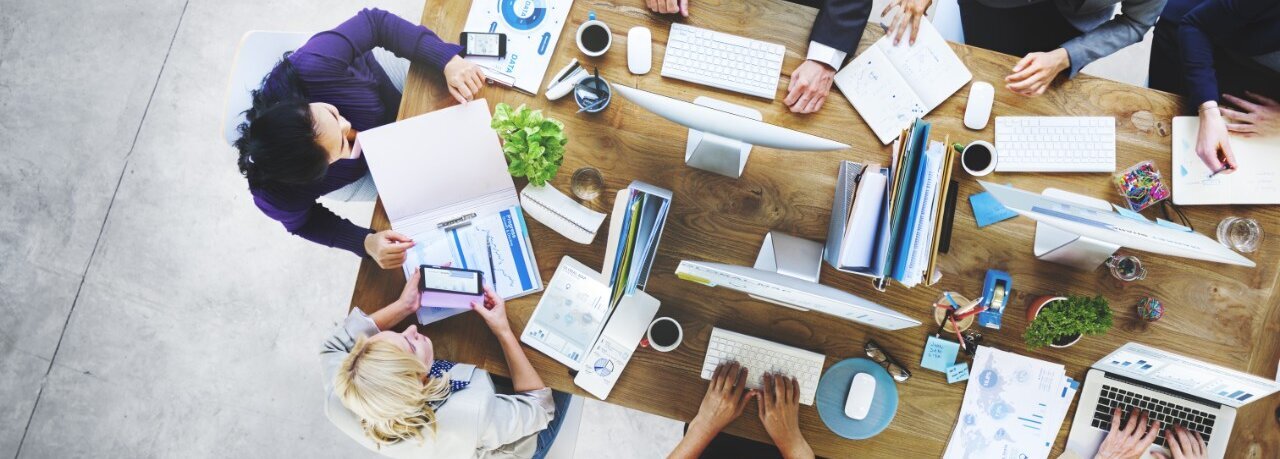 Ein großer Arbeitstisch mit mehreren arbeitenden Menschen daran von Oben