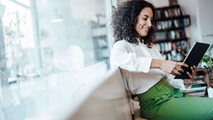 Newsletter ias aktuell Frau in weißer Bluse, grüne hose sitzt in modernem Büro und liest auf Tablett
