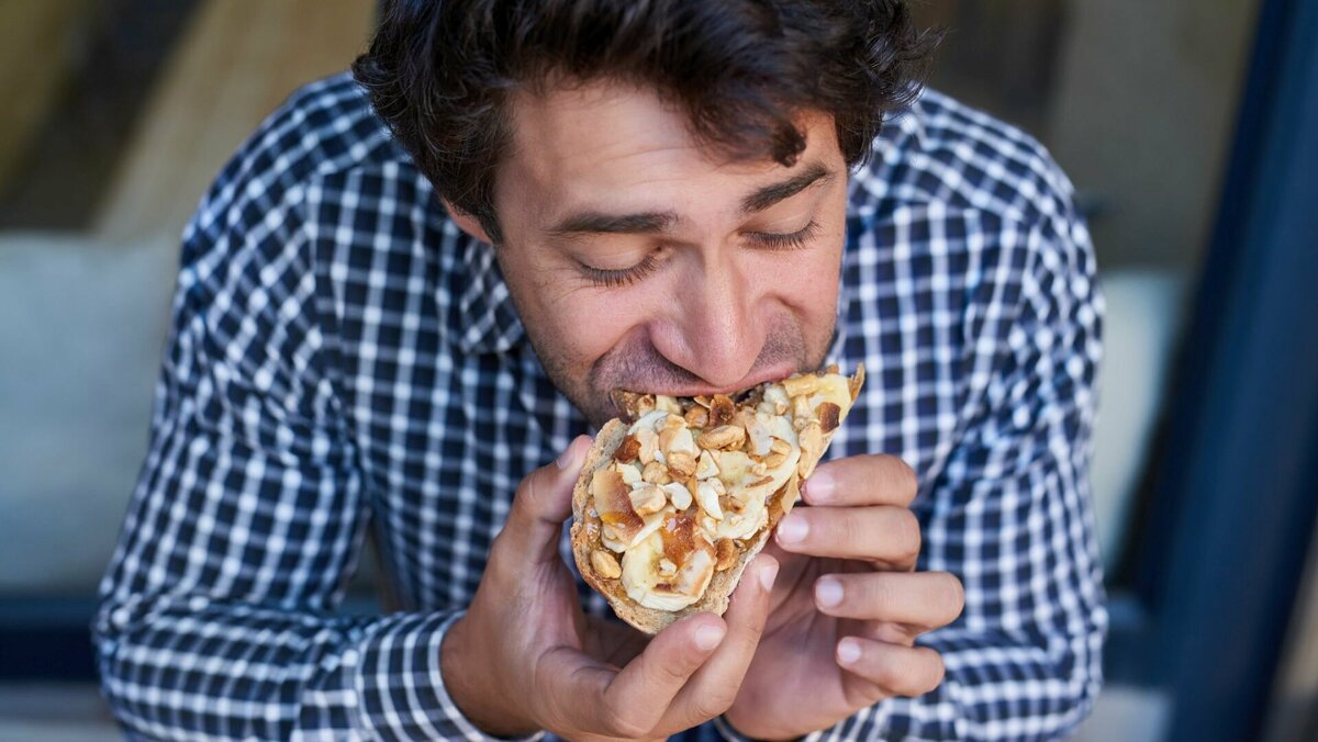 Mann im Karohemd beisst genüsslich in ein Brot mit Bananen, Honig und Nüssen