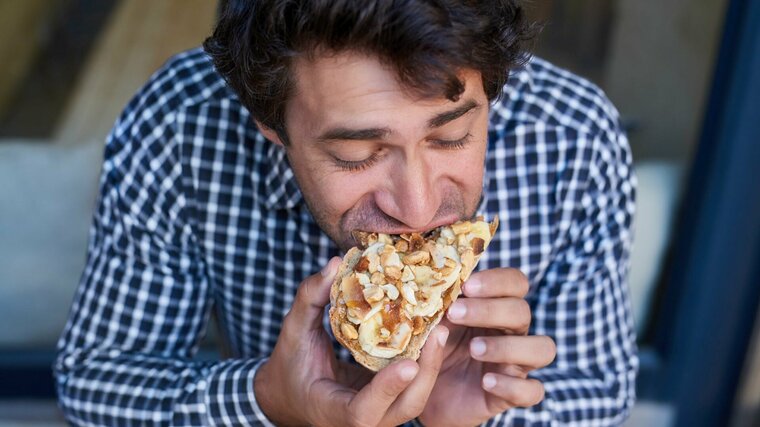 Mann im Karohemd beisst genüsslich in ein Brot mit Bananen, Honig und Nüssen