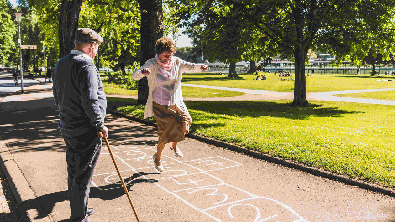Eine alte Frau springt auf einem Hinkelkästchenfeld im Park, während ihr ein alter Mann zusieht