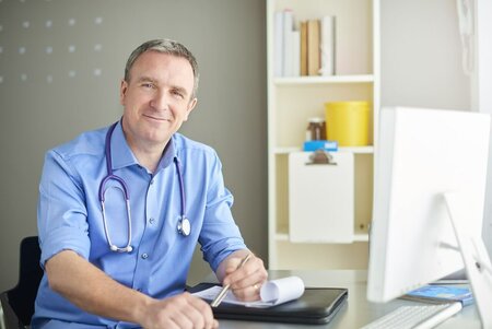 sympatisch lächelnder Arzt im blauen Hemd mit Stetoskop sitzt am Schreibtisch