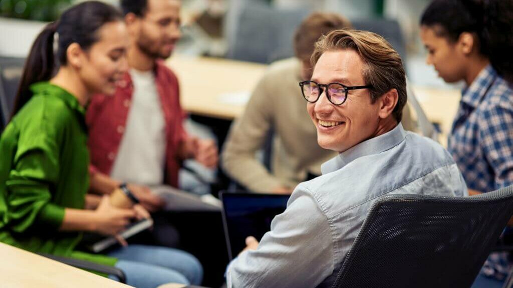 Quickcheck BEM, Betriebliches Eingliederungsmanagement lächelnder Mann mit Brille sitzend in einer Runde mit Kolleg:innen