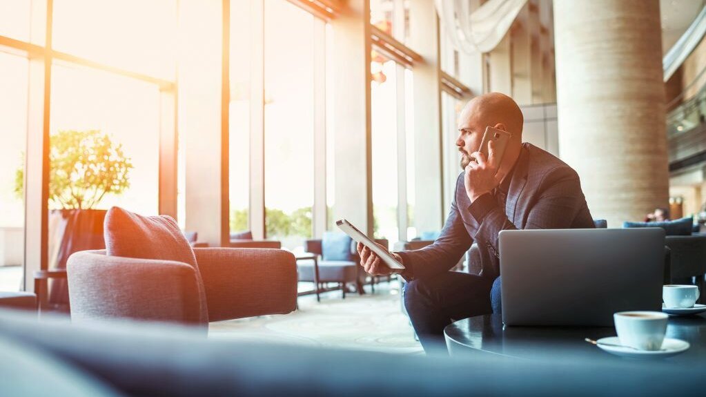 Ein Mann sitzt am Computer, hält ein Tablet in der Hand und telefoniert