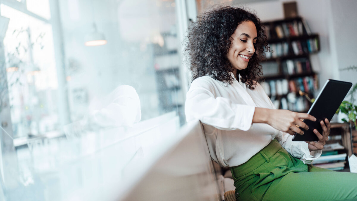 Frau in weißer Bluse, grüne hose sitzt in modernem Büro und liest auf Tablett