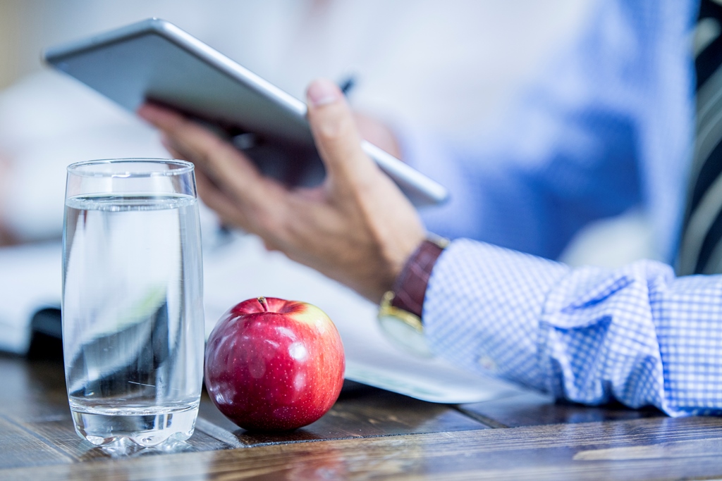Betriebliches Gesundheitsmanagement und Leistungsfähigkeitsmanagement Glas mit Wasser und einen roten Apfel im Hintergrund ein Mann mit Tablet