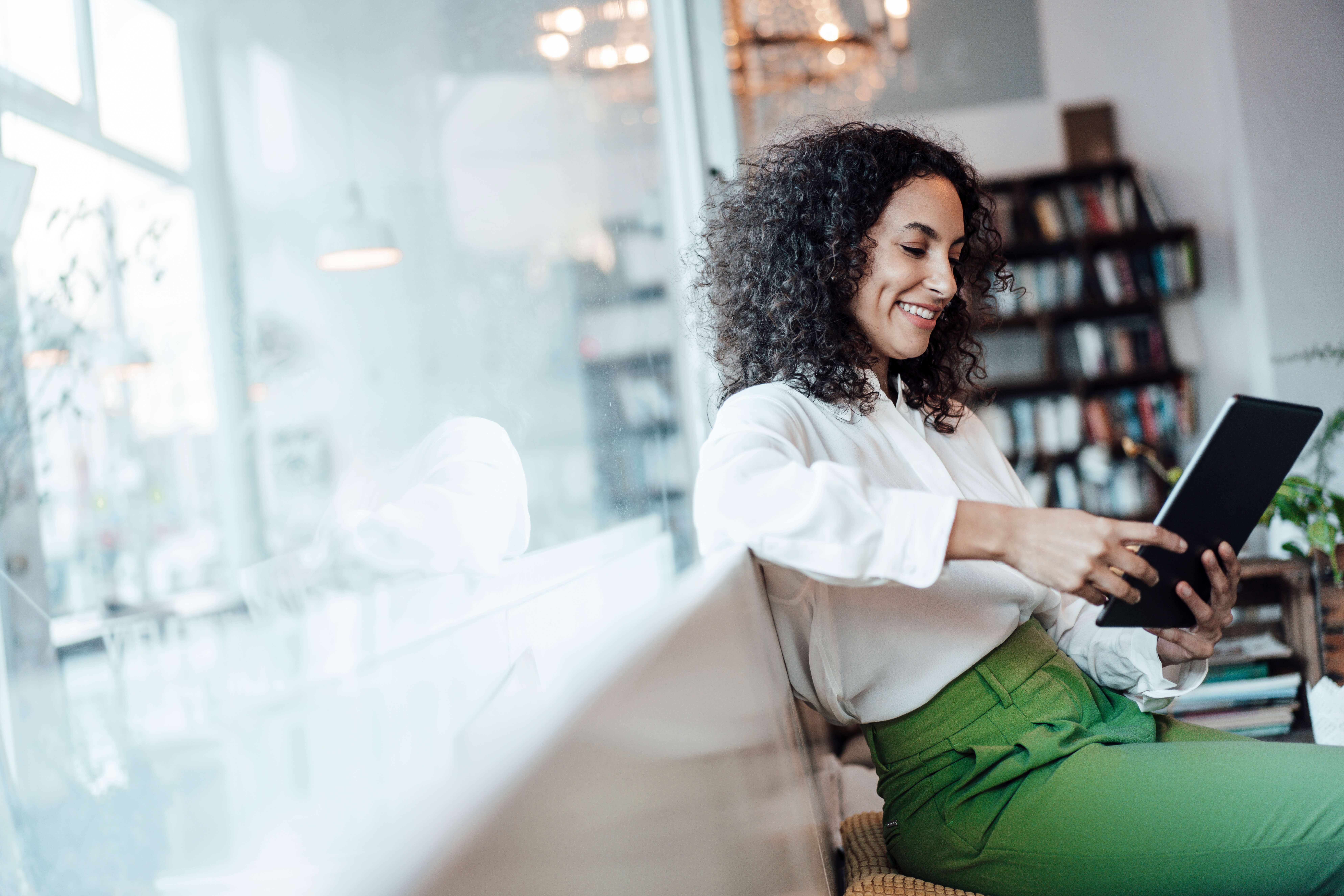 Frau in weißer Bluse, grüne hose sitzt in modernem Büro und liest auf Tablett